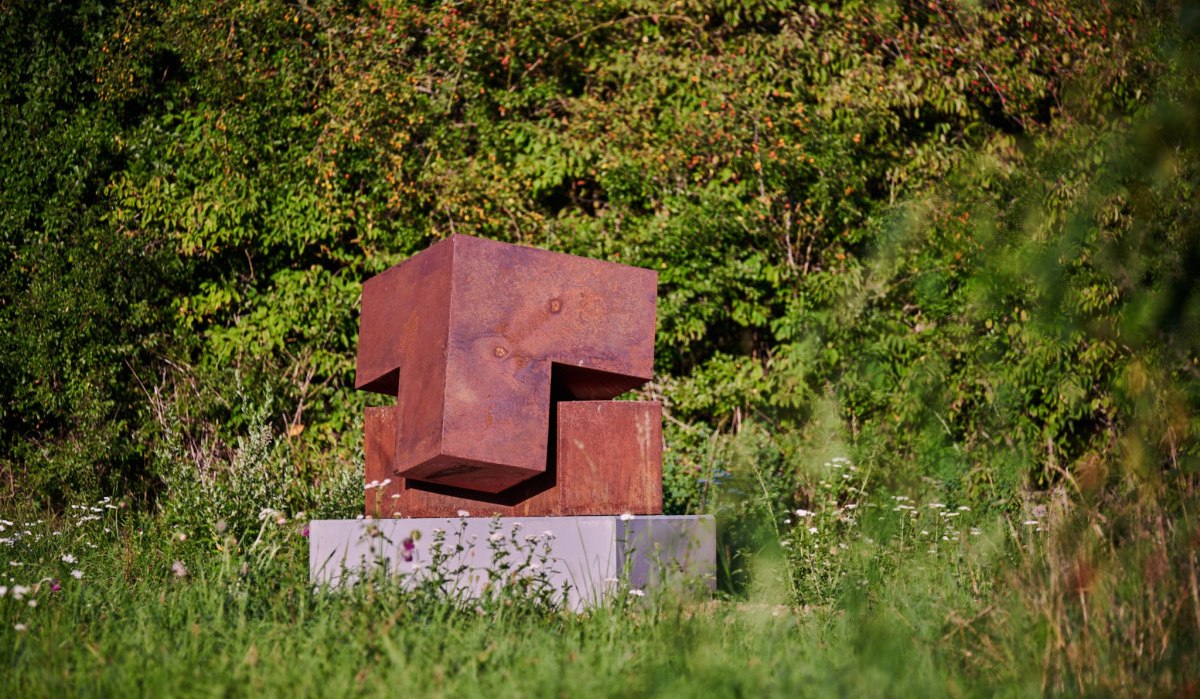 Rostige Metallskulptur auf einem Sockel, umgeben von &uuml;ppiger Vegetation und Wildblumen. Die Skulptur besteht aus geometrischen Formen., &copy; Natur.Nah. Sch&ouml;nbuch & Heckeng&auml;u