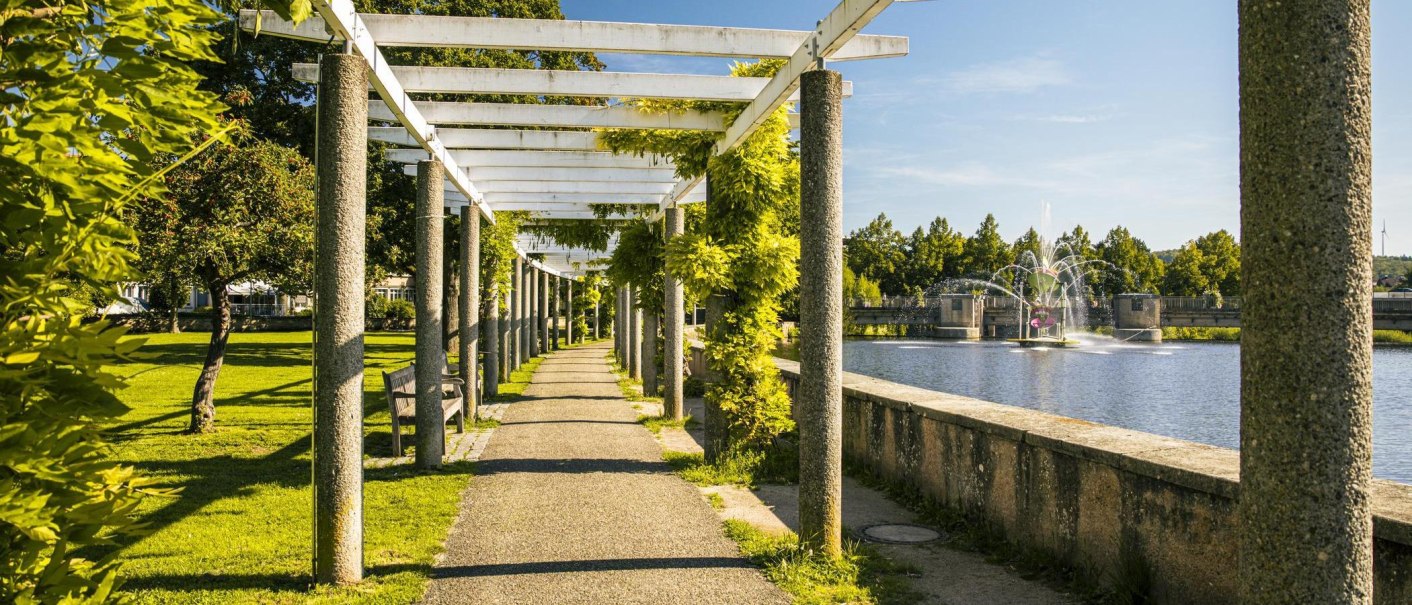Ein sonniger Weg mit Pergola und Bänken führt entlang eines künstlichen Sees mit Springbrunnen. Grüne Bäume und Pflanzen säumen den Weg., © Stuttgart-Marketing GmbH, Sarah Schmid