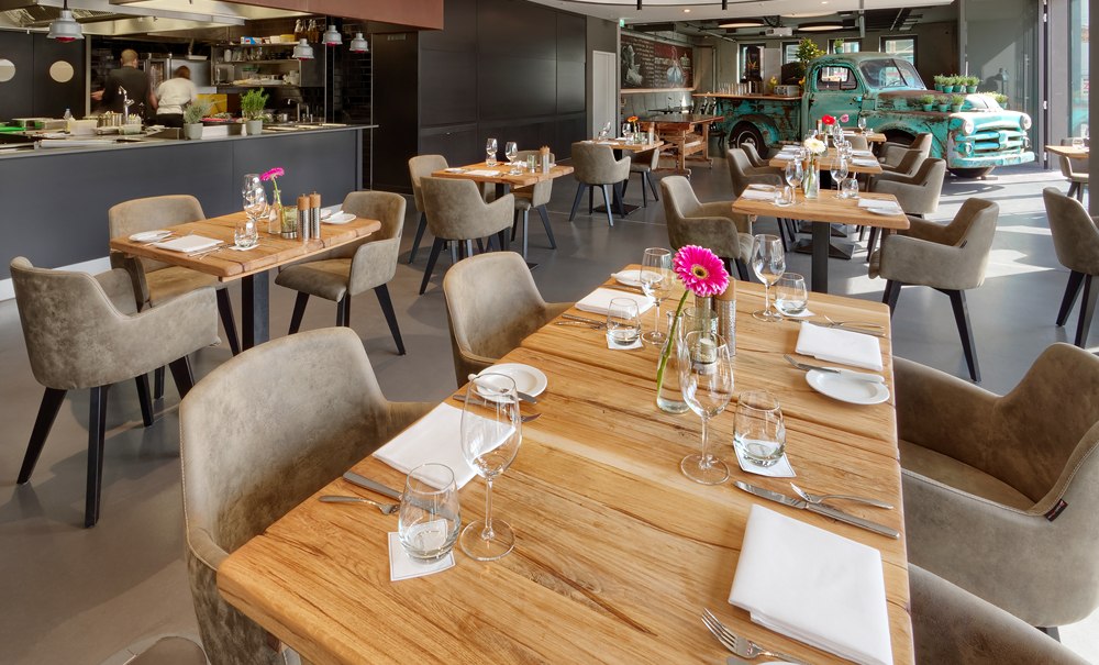 Ein modernes Restaurant mit Holztischen, stilvoll gedeckt. Im Hintergrund steht ein alter grüner Truck als Dekoration., © V8 HOTEL Motorworld Region Stuttgart Ein modernes Restaurant mit Holztischen, stilvoll gedeckt. Im Hintergrund steht ein alter grüner Truck als Dekoration., © V8 HOTEL Motorworld Region Stuttgart