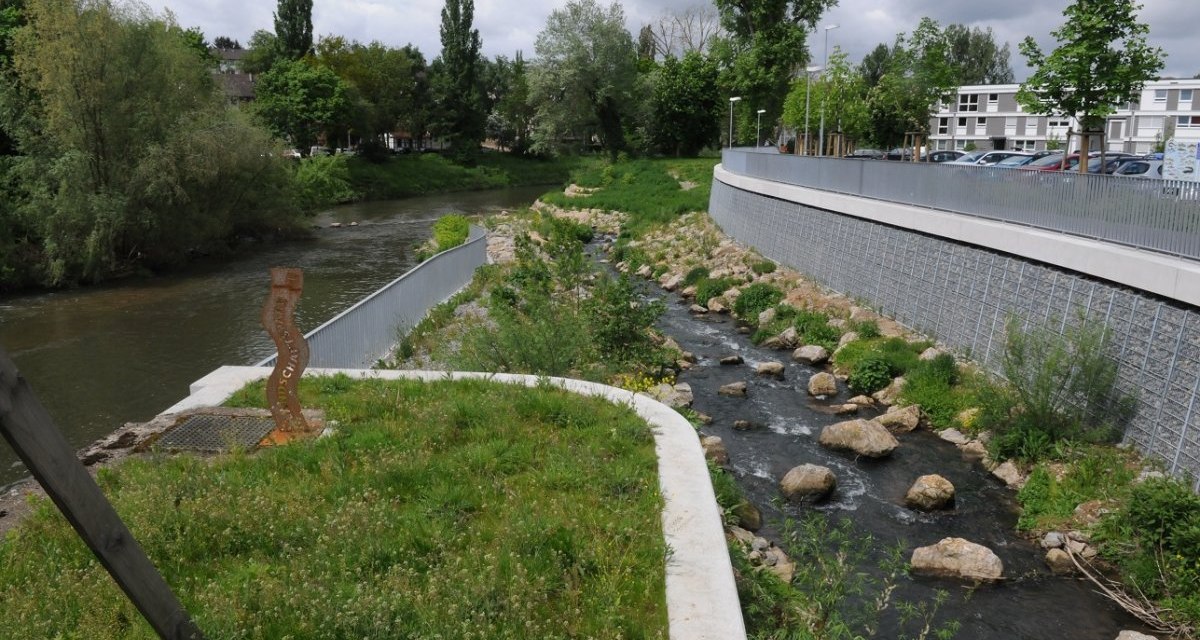 Flusslandschaft mit gr&uuml;nem Ufer, kleinem Bachlauf und B&auml;umen. Im Hintergrund sind Geb&auml;ude zu sehen., &copy; Baumg&auml;rtner