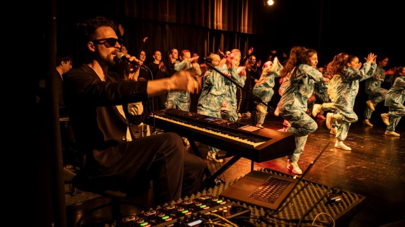 Ein Musiker mit Sonnenbrille spielt Keyboard, w&auml;hrend Kinder in blauen Kost&uuml;men auf der B&uuml;hne tanzen. Die Szene ist lebhaft und energiegeladen., &copy; PODIUM Esslingen x Kids Dance