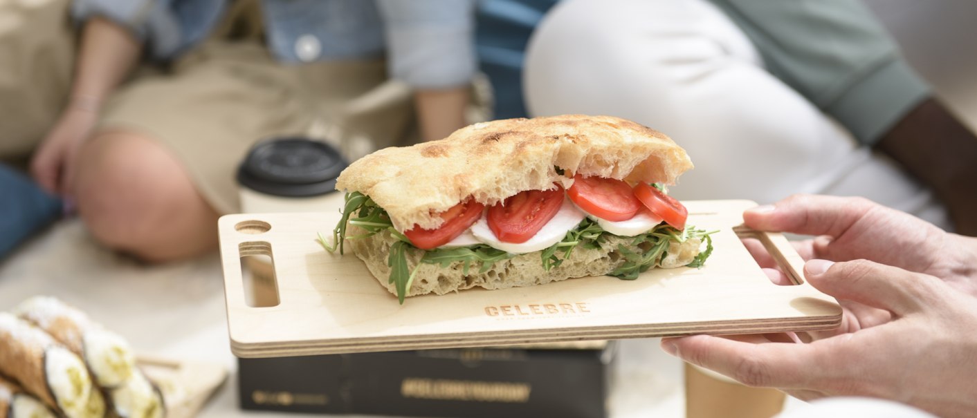 Ein Sandwich mit Tomaten, Mozzarella und Rucola auf einem Holzbrett, gehalten von einer Hand. Im Hintergrund sind Menschen bei einem Picknick zu sehen., © CELEBRE Ein Sandwich mit Tomaten, Mozzarella und Rucola auf einem Holzbrett, gehalten von einer Hand. Im Hintergrund sind Menschen bei einem Picknick zu sehen., © CELEBRE