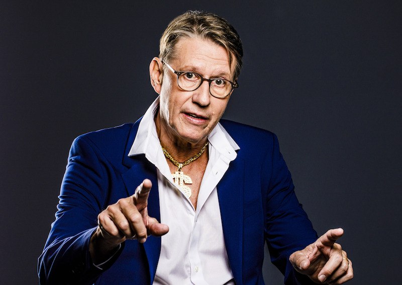 Ein Mann in einem blauen Anzug mit offenem Hemd und goldener Kette gestikuliert vor einem dunklen Hintergrund., &copy; Konzertkreis Triangel Compagnia Compostelli e.V.