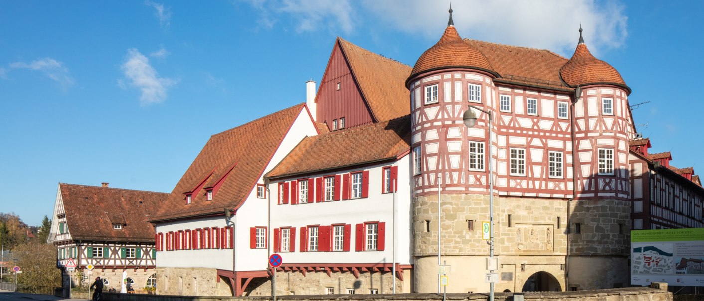 altes Schloss - rechts das Torhaus mit Sockelgeschoss aus hellem Sandstein und aufgesetztem roten Fachwerk mit weißen Gefachen, flankiert von zwei Rundtürmen. In der Bildmitte der Westflügel mit zwei aneinandergebauten, weiß verputzten Gebäudeteilen mit roten Fensterläden, überragt vom Dach und rotbraun gestrichener Giebelseite des Nordflügels. Alle Gebäudeteile eingedekct mit roten Ziegeln. Links das Fachwerkgebäude der ehemaligen Bentheimschen Forstverwaltung mit hellem Sandsteinsockel und grünen Fensterläden., © Stadt Gaildorf