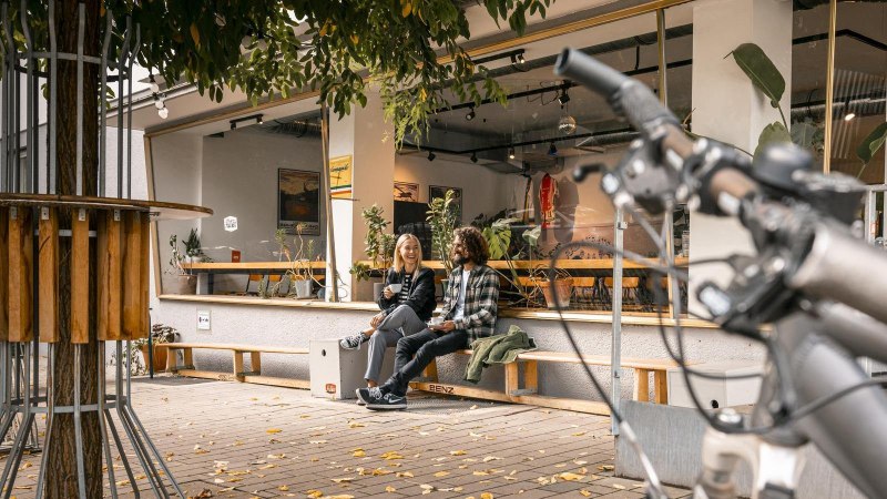 Zwei Personen sitzen lachend vor einem Caf&eacute; auf einer Bank. Im Vordergrund ist ein Fahrradlenker zu sehen. Herbstliche Bl&auml;tter liegen auf dem Boden., &copy; SMG Stuttgart Marketing GmbH - Sarah Schmid