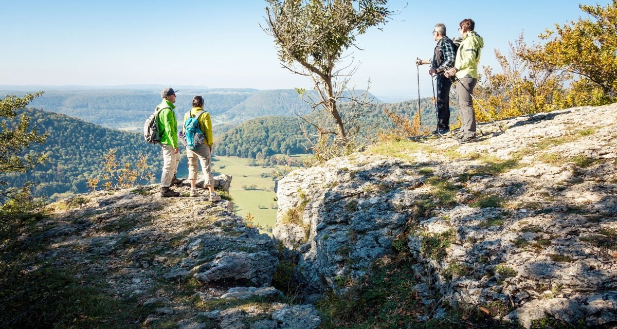 Vier Wanderer stehen auf einem Felsvorsprung am Breitenstein und genießen die Aussicht auf die bewaldete Landschaft unter einem klaren blauen Himmel., © hochgehberge Vier Wanderer stehen auf einem Felsvorsprung am Breitenstein und genießen die Aussicht auf die bewaldete Landschaft unter einem klaren blauen Himmel., © hochgehberge