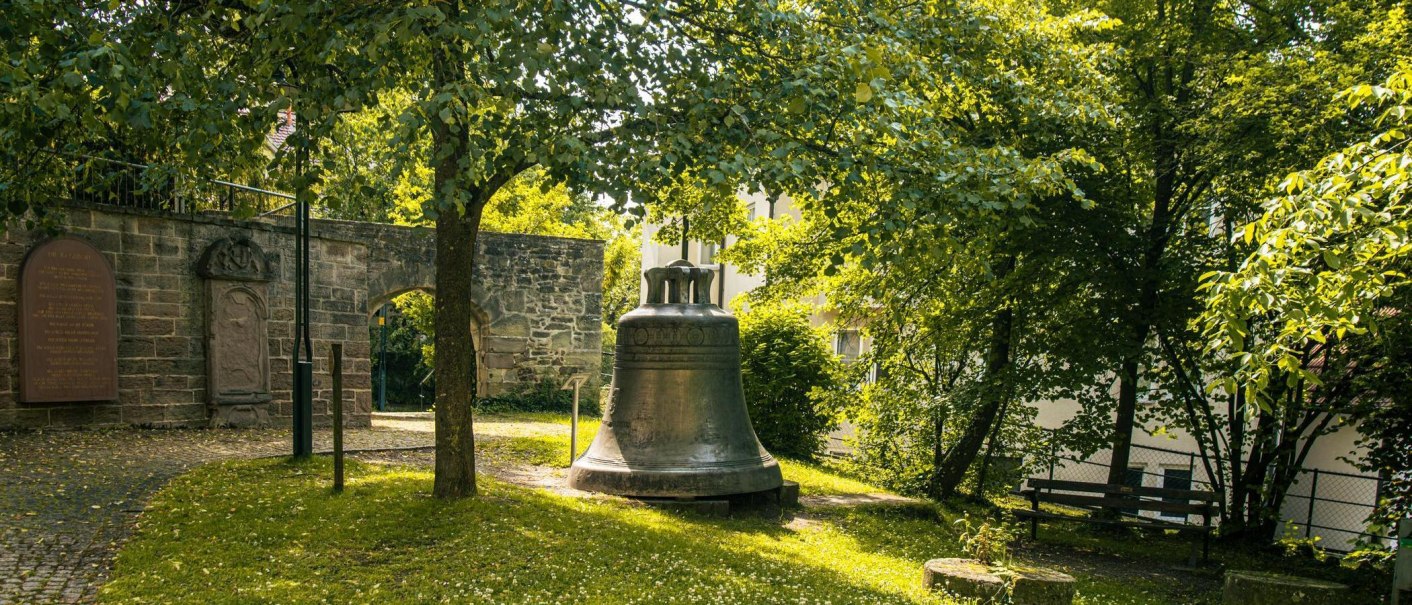 Eine große Glocke steht im Garten der Stiftskirche Herrenberg, umgeben von Bäumen und einer alten Steinmauer. Sonnenlicht fällt durch das Blätterdach., © Stuttgart-Marketing GmbH, Sarah Schmid Eine große Glocke steht im Garten der Stiftskirche Herrenberg, umgeben von Bäumen und einer alten Steinmauer. Sonnenlicht fällt durch das Blätterdach., © Stuttgart-Marketing GmbH, Sarah Schmid
