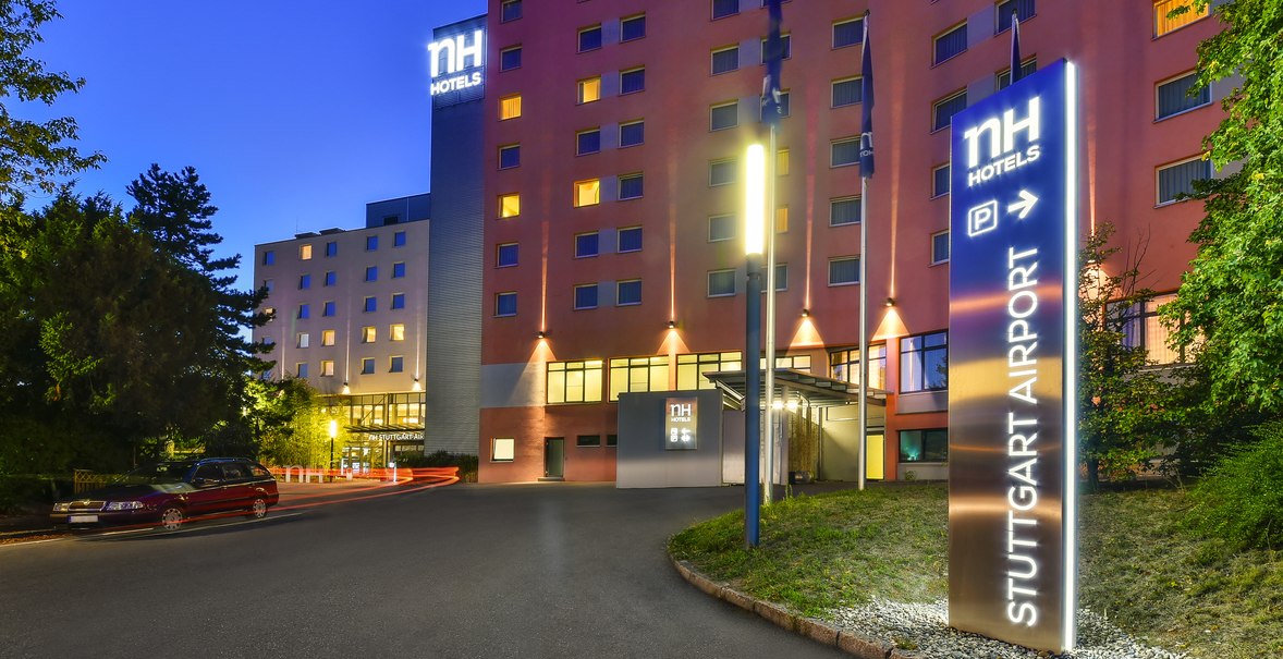Beleuchtetes NH Hotel bei Nacht mit Schild 'Stuttgart Airport'. Gebäude mit vielen Fenstern, umgeben von Bäumen., © NH Hotel Stuttgart Airport