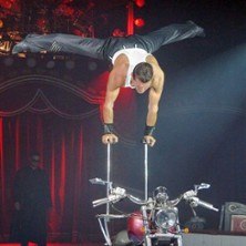 Ein Akrobat balanciert auf einer Stange über einem Motorrad im Zirkus, umgeben von rotem Bühnenlicht., © links im Bild Ein Akrobat balanciert auf einer Stange über einem Motorrad im Zirkus, umgeben von rotem Bühnenlicht., © links im Bild