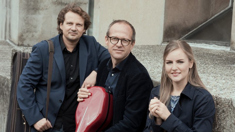 Drei Personen in eleganter Kleidung sitzen auf einer Betonmauer. Eine Person h&auml;lt ein rotes Instrumentenetui., &copy; Schwarzenberg Trio