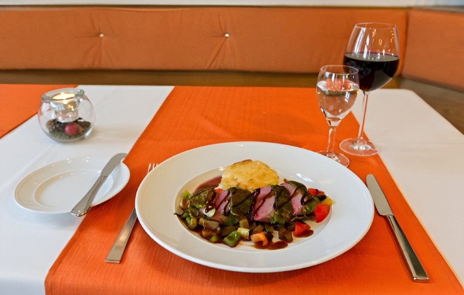 Ein Teller mit Fleisch und Gemüse auf orange-weißer Tischdecke, daneben Rotwein und Wasser. Elegante Restaurantatmosphäre., © Hotel Neuwirtshaus Ein Teller mit Fleisch und Gemüse auf orange-weißer Tischdecke, daneben Rotwein und Wasser. Elegante Restaurantatmosphäre., © Hotel Neuwirtshaus