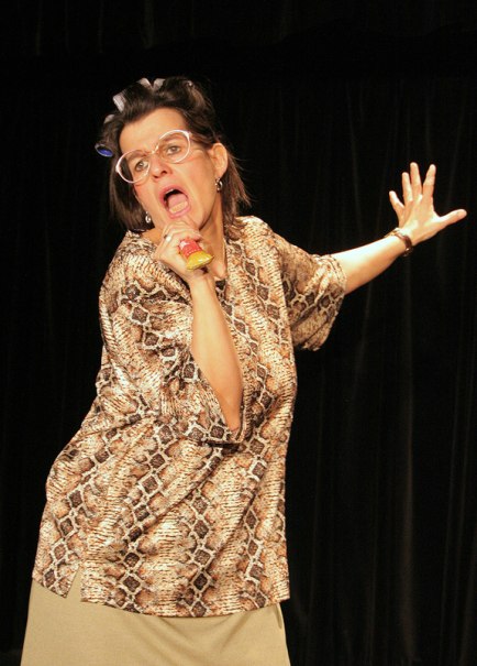 Frau mit Brille und Lockenwicklern in clownesker Pose auf Bühne, trägt gemustertes Oberteil., © Galli Theater Backnang