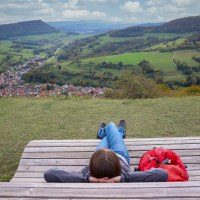 Person entspannt auf einer Holzbank mit Blick auf ein Tal, grüne Felder und ein Dorf. Ein roter Rucksack liegt daneben. Der Himmel ist bewölkt., © Foto: Mario Klaiber