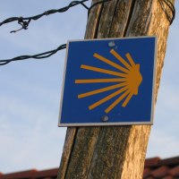 Ein blaues Schild mit gelber Jakobsmuschel als Wegzeichen des Jakobswegs, befestigt an einem Holzpfosten unter freiem Himmel., © SWT Ein blaues Schild mit gelber Jakobsmuschel als Wegzeichen des Jakobswegs, befestigt an einem Holzpfosten unter freiem Himmel., © SWT