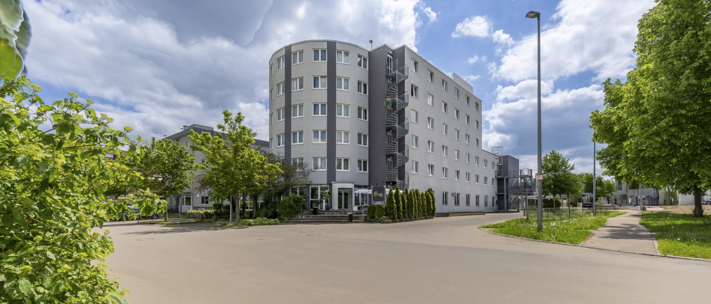 Modernes, mehrstöckiges Gebäude mit runder Ecke, grauer Fassade, umgeben von Bäumen und Gehweg, unter bewölktem Himmel., © Plaza INN Stuttgart-Filderstadt Modernes, mehrstöckiges Gebäude mit runder Ecke, grauer Fassade, umgeben von Bäumen und Gehweg, unter bewölktem Himmel., © Plaza INN Stuttgart-Filderstadt
