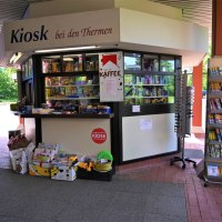 Ein Kiosk mit der Aufschrift 'Kiosk bei den Thermen', umgeben von Zeitschriftenständern und Verkaufsartikeln. Im Hintergrund sind Bäume sichtbar. Ein Kiosk mit der Aufschrift 'Kiosk bei den Thermen', umgeben von Zeitschriftenständern und Verkaufsartikeln. Im Hintergrund sind Bäume sichtbar.