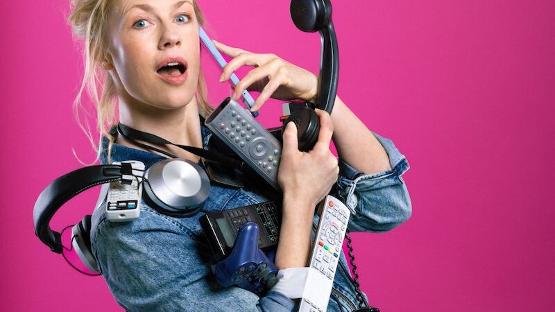 Frau in Jeansjacke hält viele elektronische Geräte wie Fernbedienungen und Kopfhörer vor einem pinken Hintergrund., © Mauerwerk GmbH