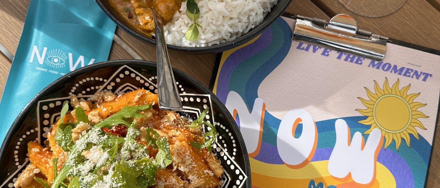 Ein Tisch mit Curry und Reis, Pasta mit Rucola, einem Getr&auml;nk und einer bunten Speisekarte mit der Aufschrift 'NOW'., &copy; cNOW