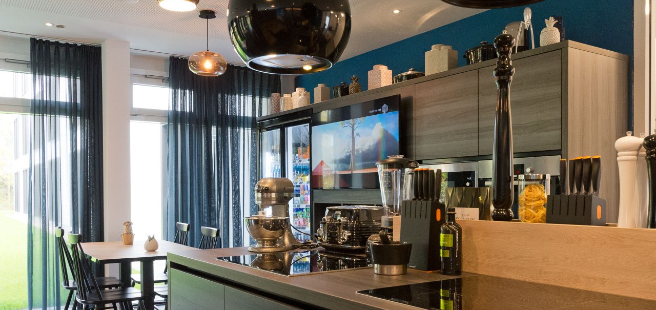 Moderne Küche mit Kochinsel, Esstisch, Hängelampen und Geräten. Große Fenster mit blauen Vorhängen lassen viel Licht herein., © Arthotel Ana / elaya