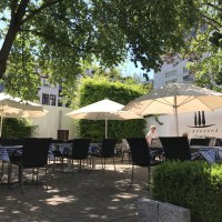 Ein sonniger Garten mit Tischen, Stühlen und Sonnenschirmen, umgeben von Bäumen und Pflanzen. Im Hintergrund ist eine Wand mit der Aufschrift 'Il Boccone' sichtbar., © Restaurant Il Boccone Ein sonniger Garten mit Tischen, Stühlen und Sonnenschirmen, umgeben von Bäumen und Pflanzen. Im Hintergrund ist eine Wand mit der Aufschrift 'Il Boccone' sichtbar., © Restaurant Il Boccone