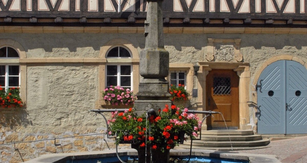 Ein steinerner Brunnen mit Blumen steht vor einem historischen Fachwerkhaus mit blauen Fensterl&auml;den und einem Holztor., &copy; Natur.Nah. Sch&ouml;nbuch & Heckeng&auml;u