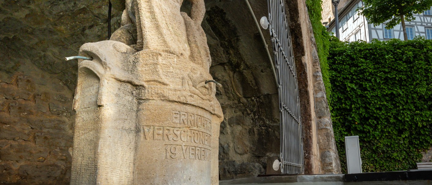 Steinfigur am Marktbrunnen in Backnang, darunter Inschrift. Im Hintergrund ein Turm mit Uhr und Fachwerk. Wasserbecken im Vordergrund., © René Straube