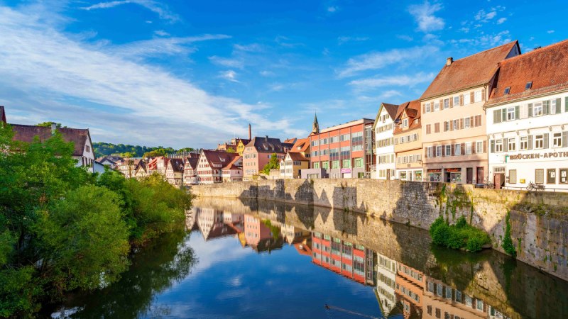Fachwerkhäuser und moderne Gebäude spiegeln sich im Fluss in Schwäbisch Hall. Klare blaue Himmel und grüne Vegetation rahmen die Szene ein., © Michael Kühneisen Fachwerkhäuser und moderne Gebäude spiegeln sich im Fluss in Schwäbisch Hall. Klare blaue Himmel und grüne Vegetation rahmen die Szene ein., © Michael Kühneisen