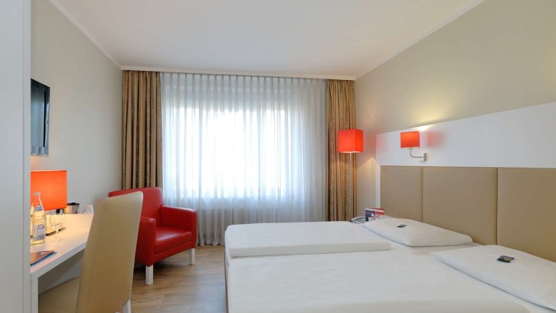 Modernes Hotelzimmer mit zwei Einzelbetten, rotem Sessel, Schreibtisch und Fenster mit Vorh&auml;ngen. Rote Lampen sorgen f&uuml;r Akzente., &copy; Hotel Stuttgart-Sindelfingen City by Tulip Inn