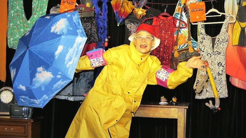 Eine Person in gelbem Regenmantel und rotem Hut posiert mit einem blauen Regenschirm vor einer bunten Kulisse aus Kleidung und Accessoires., &copy; Galli Theater Backnang