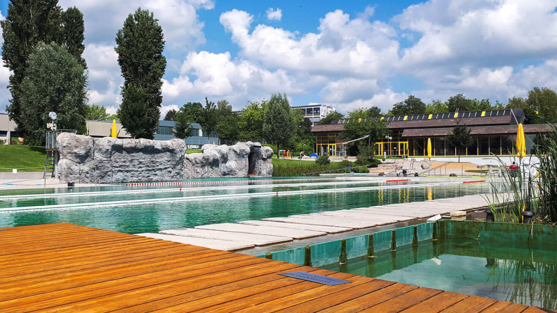 Das Naturfreibad Herrenberg zeigt einen Pool mit Holzdeck, Felsenwand und umgebender grüner Landschaft unter blauem Himmel mit Wolken., © Stadtwerke Herrenberg Das Naturfreibad Herrenberg zeigt einen Pool mit Holzdeck, Felsenwand und umgebender grüner Landschaft unter blauem Himmel mit Wolken., © Stadtwerke Herrenberg