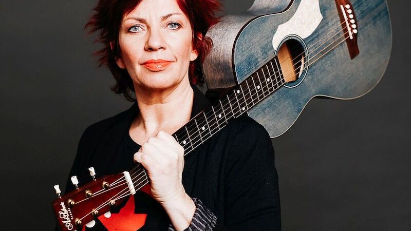 Eine Person mit rotem Haar hält eine blaue Gitarre über der Schulter und schaut direkt in die Kamera. Der Hintergrund ist dunkel., © Stadthalle Leonberg