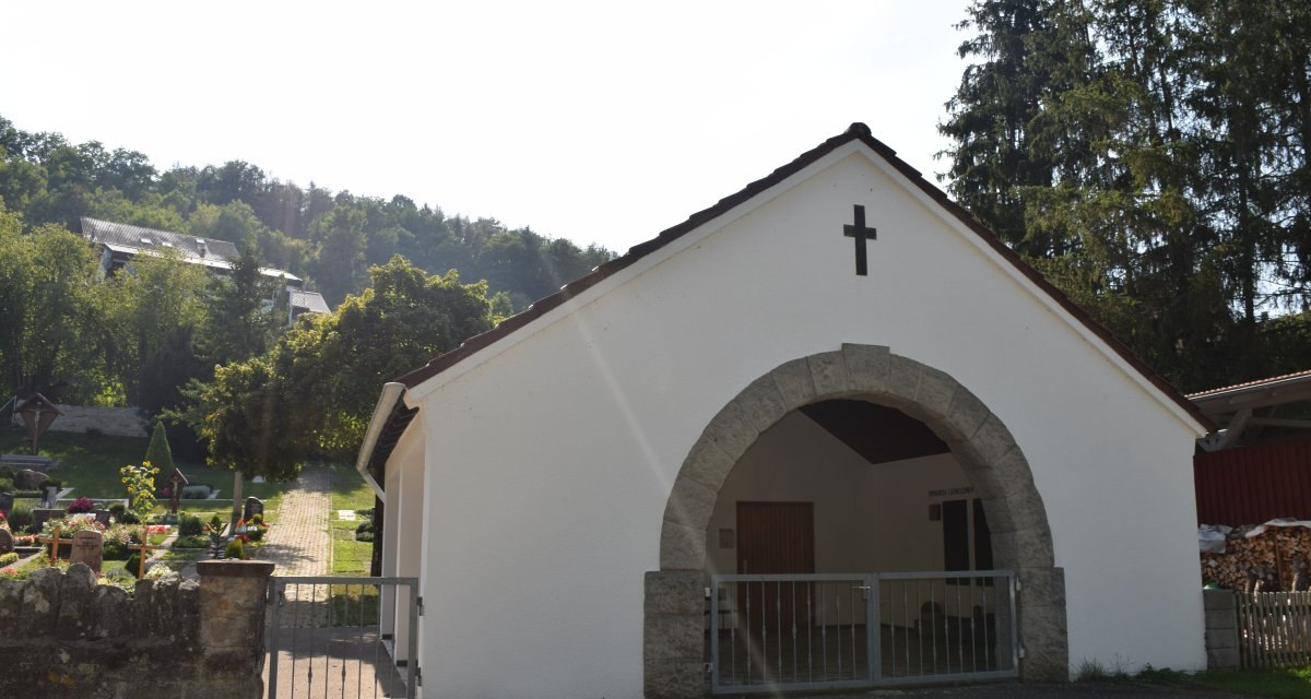 Wei&szlig;e Kapelle mit Kreuz, umgeben von B&auml;umen und einem Friedhof. Ein Weg f&uuml;hrt durch das Gel&auml;nde. Im Hintergrund sind H&uuml;gel und Geb&auml;ude zu sehen., &copy; Land der 1000 H&uuml;gel - Kraichgau-Stromberg