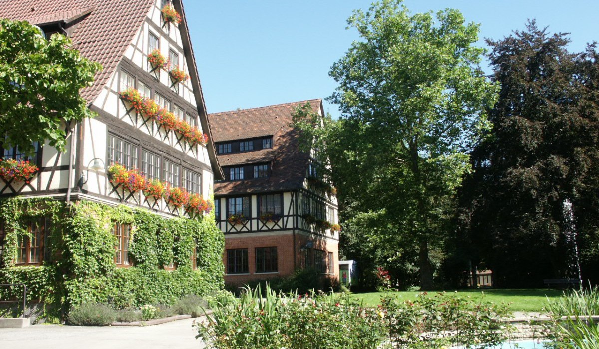Fachwerkhaus mit blühenden Blumenkästen und grünem Efeu. Im Vordergrund ein gepflegter Garten mit Brunnen, umgeben von Bäumen., © Natur.Nah. Schönbuch & Heckengäu Fachwerkhaus mit blühenden Blumenkästen und grünem Efeu. Im Vordergrund ein gepflegter Garten mit Brunnen, umgeben von Bäumen., © Natur.Nah. Schönbuch & Heckengäu