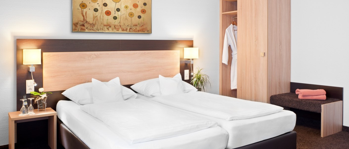 Ein modernes Hotelzimmer mit Doppelbett, Nachttisch, Schrank und Wandbild. Ein Bademantel hängt im Schrank, Handtücher liegen auf einer Bank., © TOMAS