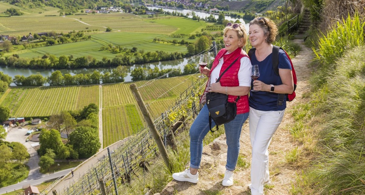 Zwei Frauen wandern mit Weingläsern durch Weinberge. Im Hintergrund sind ein Fluss und grüne Felder zu sehen.