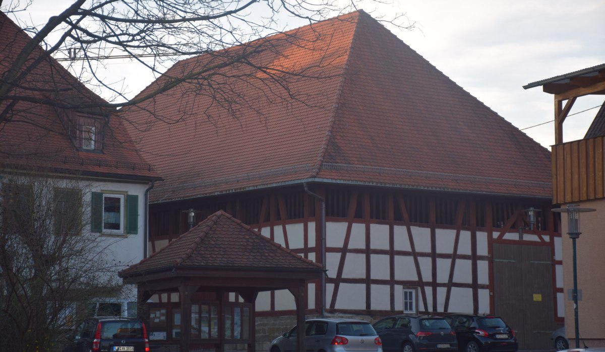Ein großes Fachwerkgebäude mit rotem Ziegeldach, umgeben von geparkten Autos und einem Baum im Vordergrund., © Land der 1000 Hügel - Kraichgau-Stromberg Ein großes Fachwerkgebäude mit rotem Ziegeldach, umgeben von geparkten Autos und einem Baum im Vordergrund., © Land der 1000 Hügel - Kraichgau-Stromberg