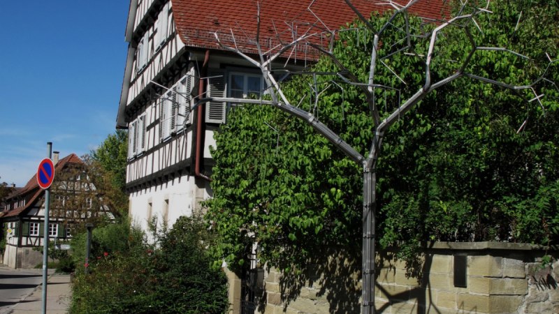 Ein Brunnen mit einer Baumskulptur aus Metall vor einem Fachwerkhaus. Der Himmel ist klar und blau, umgeben von grüner Vegetation., © RadL Leonberg Ein Brunnen mit einer Baumskulptur aus Metall vor einem Fachwerkhaus. Der Himmel ist klar und blau, umgeben von grüner Vegetation., © RadL Leonberg