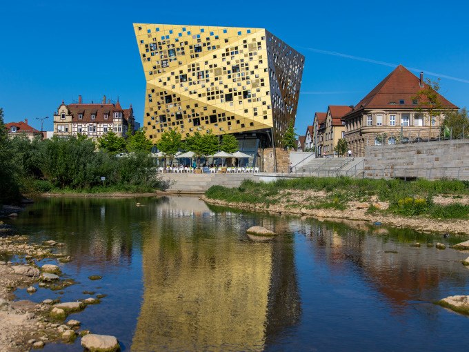 Ein modernes, goldenes Gebäude spiegelt sich in einem Fluss. Historische Gebäude und grüne Uferpflanzen umgeben die Szene unter klarem, blauem Himmel., © Touristik und Marketing GmbH Ein modernes, goldenes Gebäude spiegelt sich in einem Fluss. Historische Gebäude und grüne Uferpflanzen umgeben die Szene unter klarem, blauem Himmel., © Touristik und Marketing GmbH