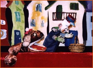 Kasperltheater-Szene mit einer Schlange und einer Figur vor bunten H&auml;usern. Die Figuren interagieren auf einer B&uuml;hne mit rotem Vorhang., &copy; Bildrechte: Gregor Oehmann