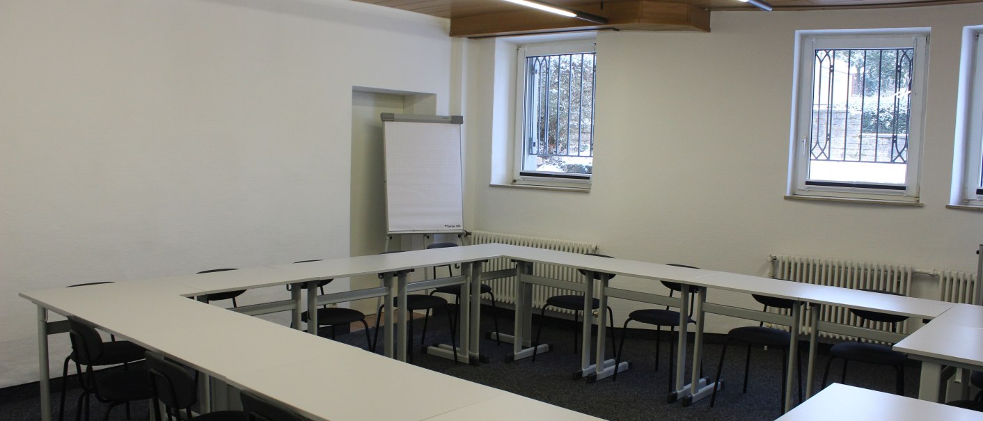Ein leerer Konferenzraum mit U-förmiger Tischanordnung, mehreren Stühlen und einem Flipchart an der Wand., © TOMAS Ein leerer Konferenzraum mit U-förmiger Tischanordnung, mehreren Stühlen und einem Flipchart an der Wand., © TOMAS