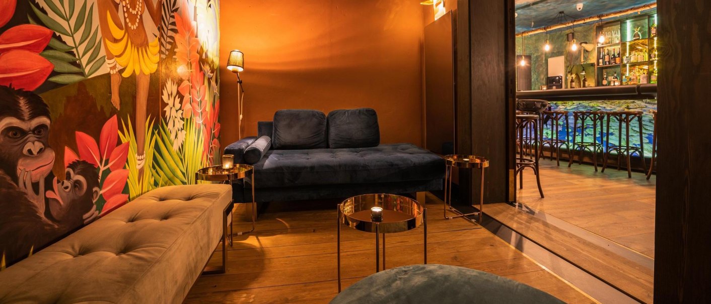 Gemütliche Lounge mit blauer Couch, buntem Wandbild mit Affen und Pflanzen. Im Hintergrund eine Bar mit beleuchteten Regalen und Hockern., © Holzmaler, Stuttgart