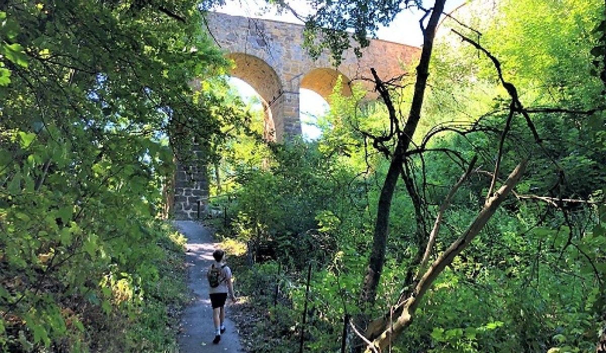 Ein Wanderer auf einem schattigen Pfad unter einer steinernen Bogenbrücke, umgeben von dichtem Grün., © Touristik und Marketing GmbH Ein Wanderer auf einem schattigen Pfad unter einer steinernen Bogenbrücke, umgeben von dichtem Grün., © Touristik und Marketing GmbH