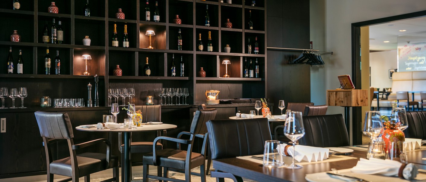 Elegantes Restaurant mit dunklen Holzm&ouml;beln, gedeckten Tischen und einem gro&szlig;en Regal voller Weinflaschen und Gl&auml;ser., &copy; ARCOTEL Hotels