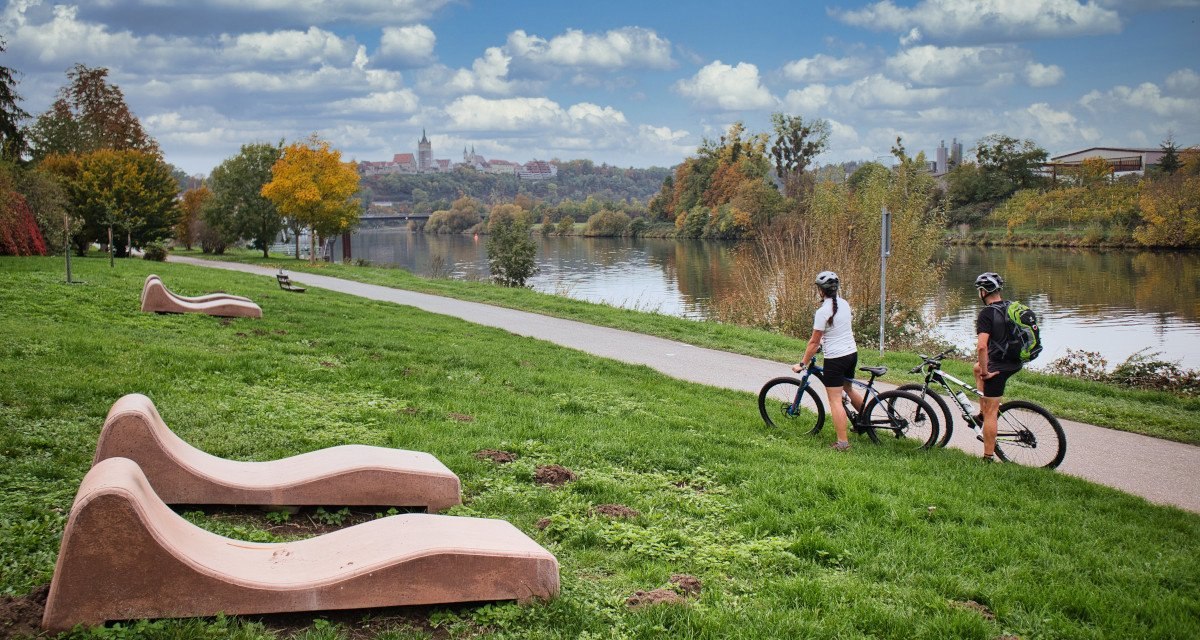 Zwei Radfahrer auf einem Weg am Neckartal-Radweg bei Bad Wimpfen, umgeben von gr&uuml;ner Landschaft und Fluss. Im Hintergrund ist die Stadt sichtbar., &copy; Gesch&auml;ftsstelle Neckartal-Radweg Adrian Weng
