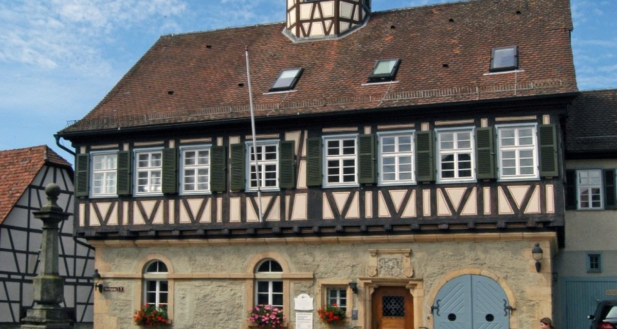 Fachwerkgeb&auml;ude mit Turm und Brunnen auf gepflastertem Platz, blauer Himmel im Hintergrund., &copy; Natur.Nah. Sch&ouml;nbuch & Heckeng&auml;u