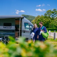 Ein Paar in blauen Badem&auml;nteln spaziert neben einem geparkten Wohnmobil auf einem sonnigen Campingplatz. Im Vordergrund stehen ein Tisch und St&uuml;hle., &copy; Thomas Niederm&uuml;ller