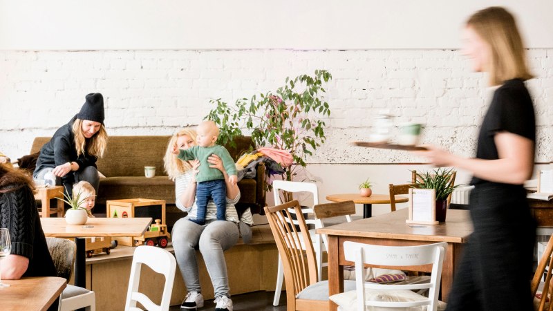 Gemütliches Café mit spielenden Kindern und Erwachsenen. Eine Bedienung serviert Getränke. Pflanzen und Holzmöbel schaffen eine einladende Atmosphäre., © Sven Weber Gemütliches Café mit spielenden Kindern und Erwachsenen. Eine Bedienung serviert Getränke. Pflanzen und Holzmöbel schaffen eine einladende Atmosphäre., © Sven Weber