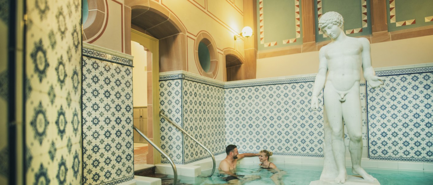 Ein kleines Becken mit blauen Fliesen und einer Statue. Zwei Personen entspannen im Wasser und genie&szlig;en die Atmosph&auml;re., &copy; &copy;LOCHER Fotodesign & Manufaktur &copy;Staatsbad Wildbad GmbH