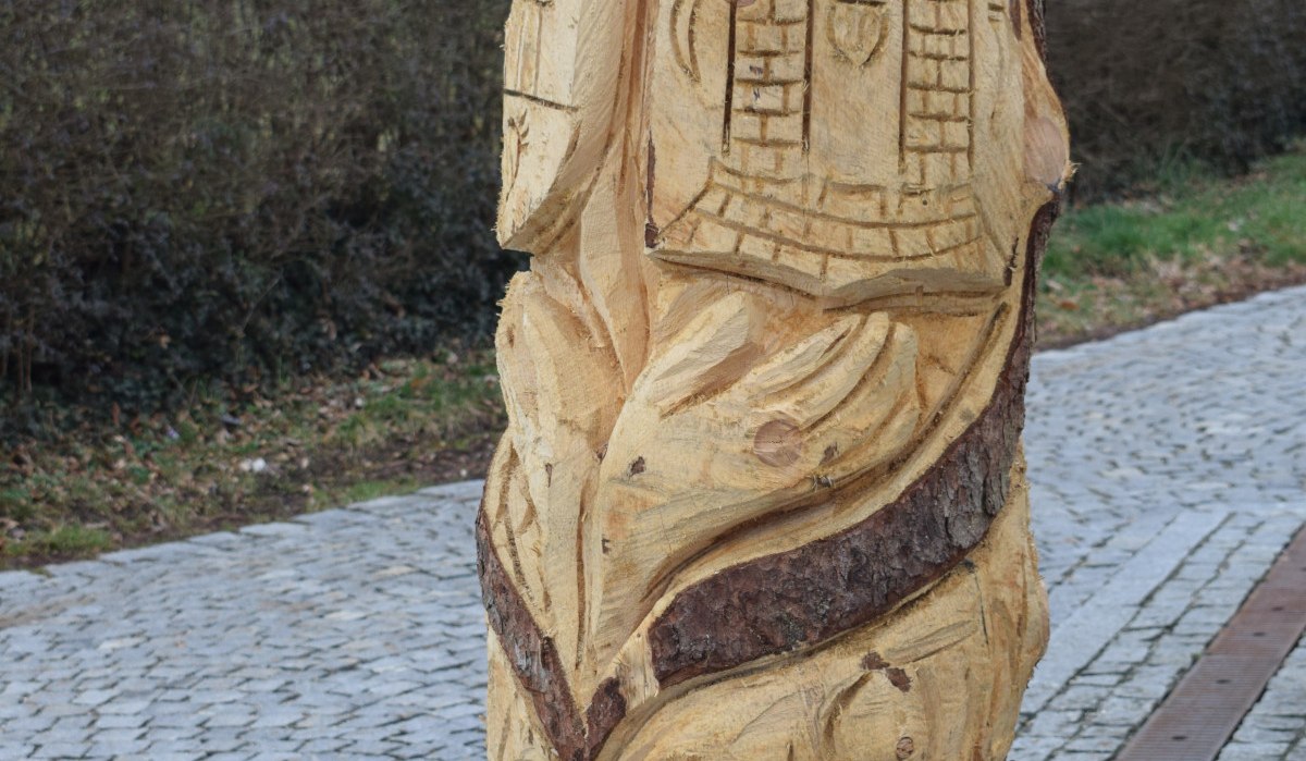 Holzskulptur mit geschnitzten Motiven, darunter ein Gesicht und Gebäude, auf einem gepflasterten Weg am Skulpturenpfad Böbingen., © Gemeinde Böbingen