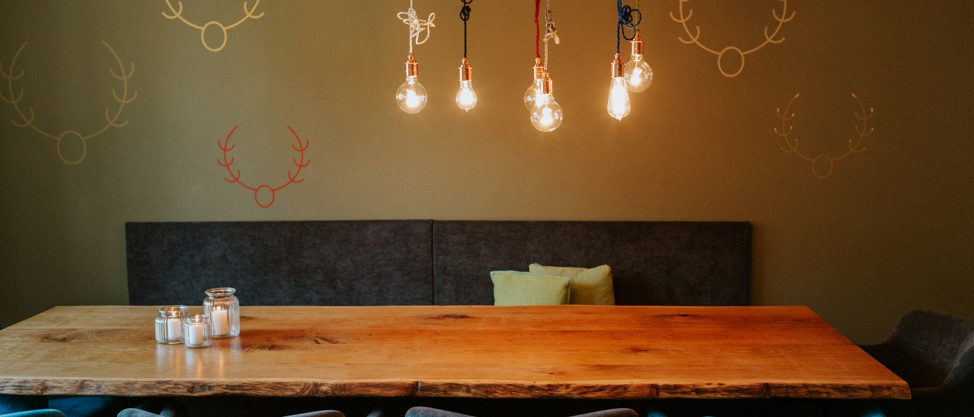 Ein langer Holztisch mit blauen Stühlen in einem Gastraum. An der Wand sind Hirschgeweihe gemalt, Glühbirnen hängen von der Decke., © Daniela Reske Ein langer Holztisch mit blauen Stühlen in einem Gastraum. An der Wand sind Hirschgeweihe gemalt, Glühbirnen hängen von der Decke., © Daniela Reske